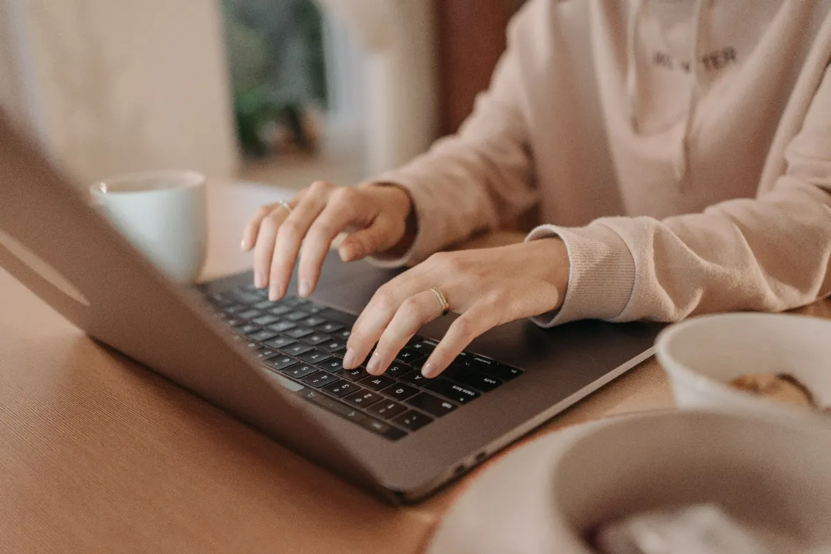 A person typing on a laptop at a wooden desk beside a mug of coffee